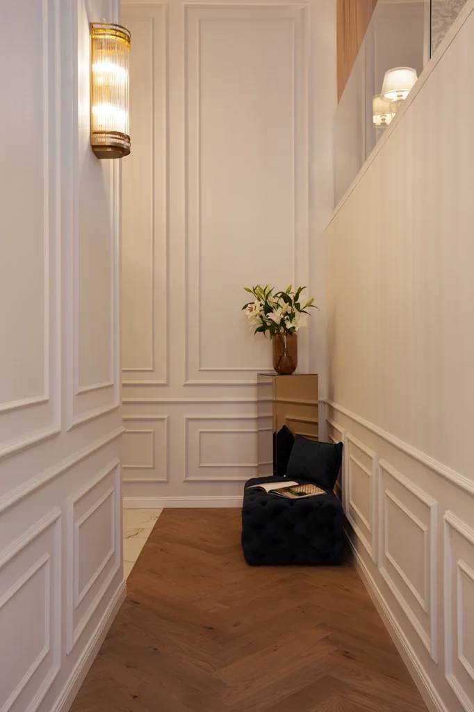 Refined transitional hallway with white panel molding, herringbone wood flooring, brass wall sconce, mirrored pedestal and MA Furniture tufted ottoman seating.