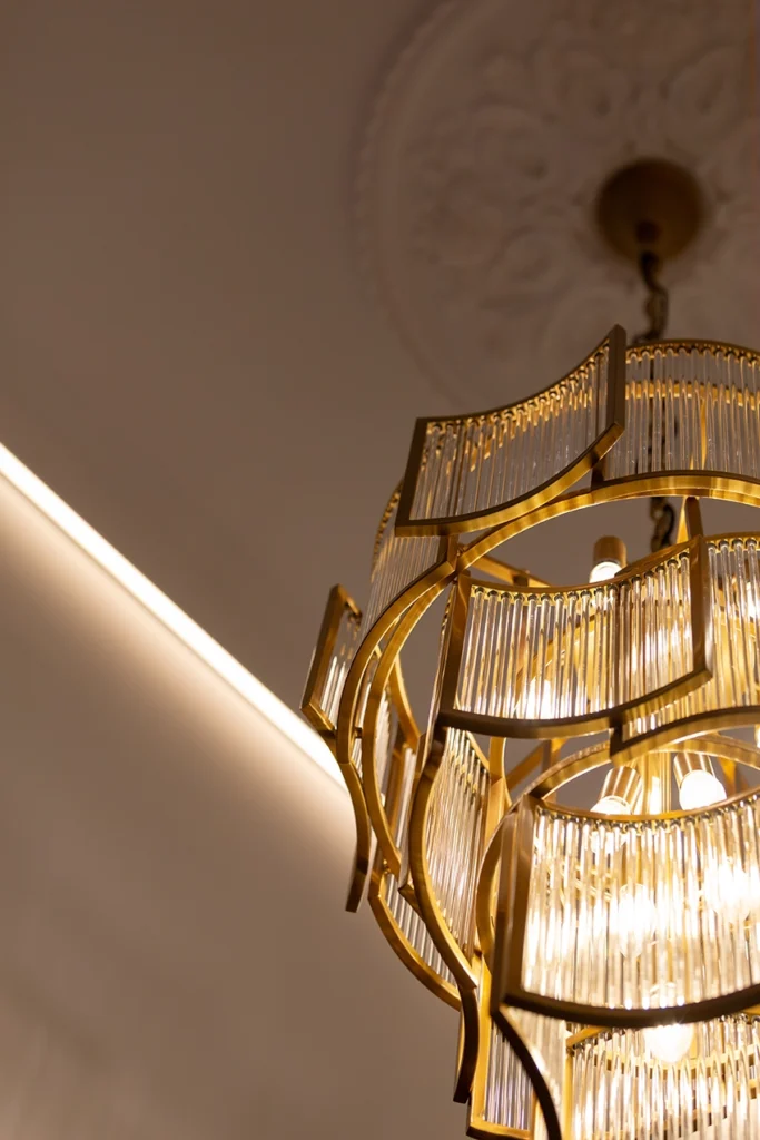 Neoclassical gold chandelier with ribbed glass panels, rosette ornate ceiling plaster work, and ambient recessed lighting.