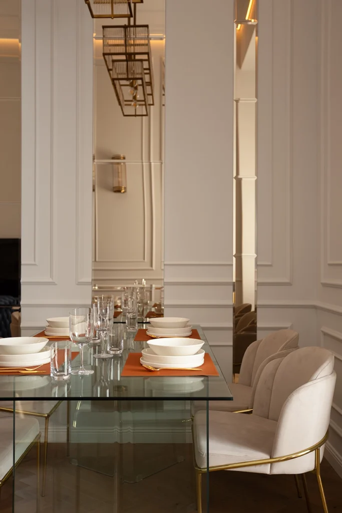 Contemporary dining room with glass table, velvet chairs with gold metal frame, mirrored wall panels, and geometric brass chandelier.
