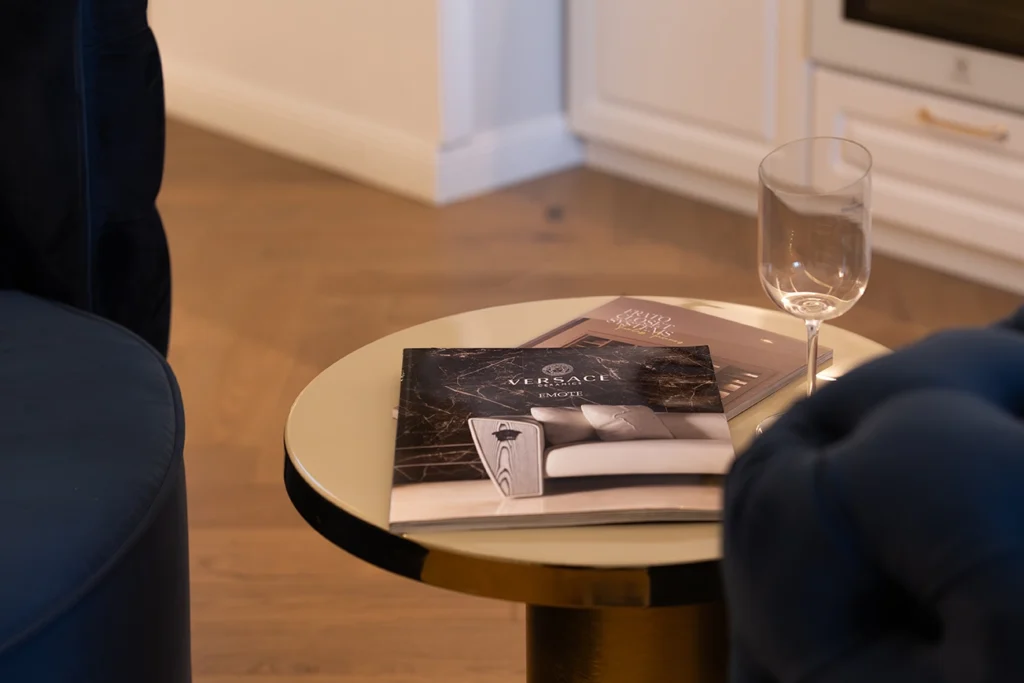 Round brass side table styled with Versace design books and a Bordeaux wine glass beside a velvet navy armchair.
