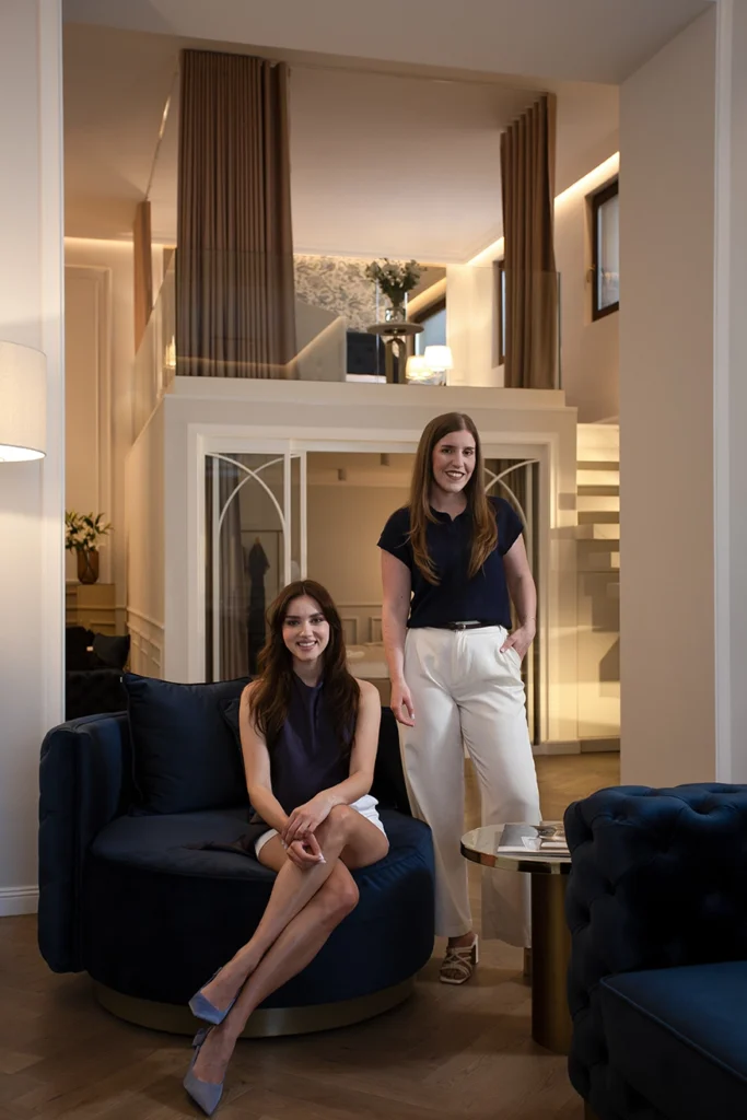 Portrait of designers Alina Ardelean and Mihaela Iuhas in a luxury modern interior with velvet navy armchair, glass mezzanine, wood paneling, and minimalist staircase.