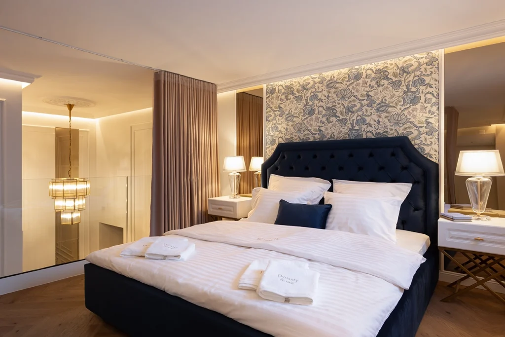 Neatly styled bed with navy upholstered headboard, crisp white linens, glass lamps, layered pillows, and folded towels in a refined hotel-inspired setting