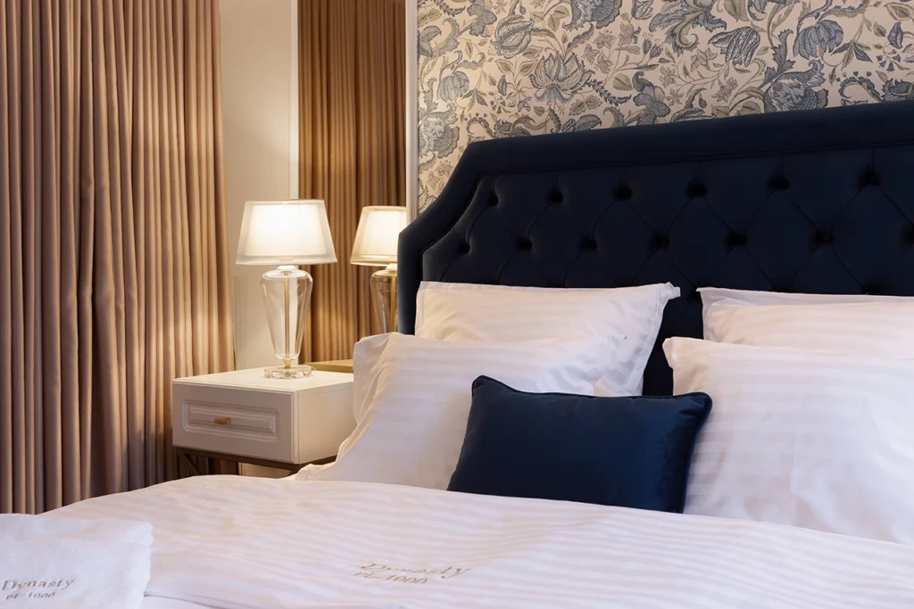 Navy tufted upholstered bed with white linens, navy accent pillow, floral wallpaper, and symmetrical nightstand styling and a glass lamp.
