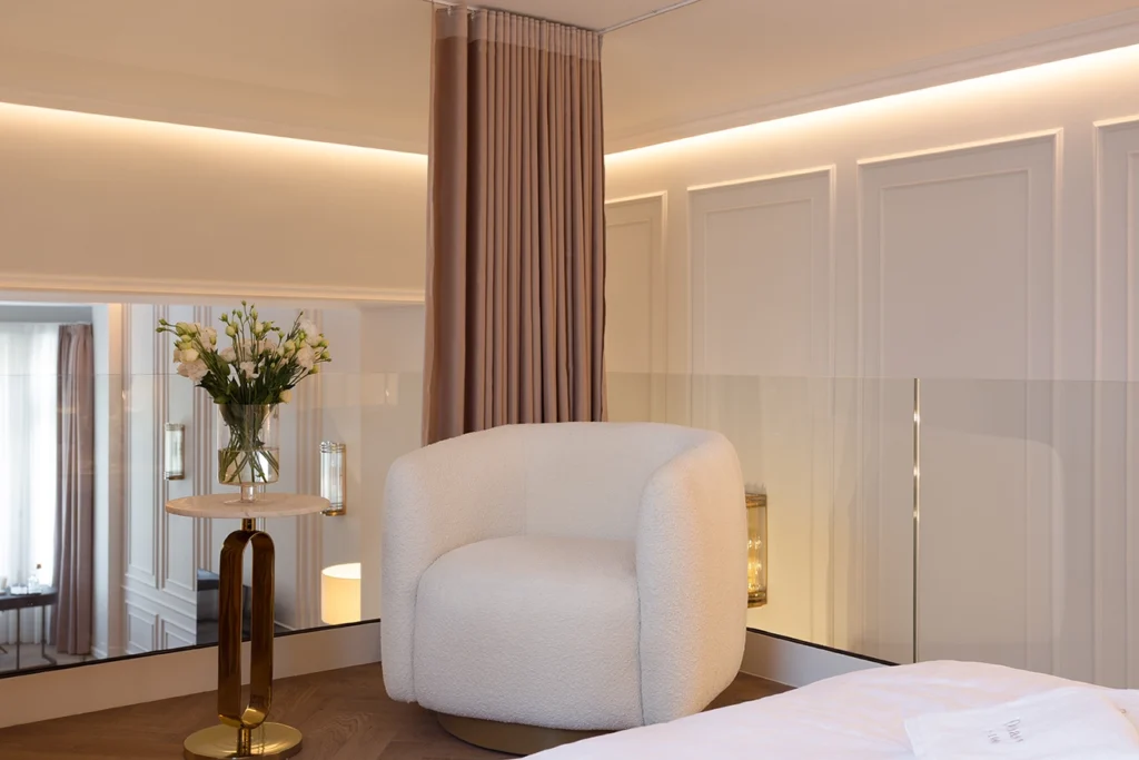 Bedroom lounge with rounded bouclé or faux-sherpa white armchair, gold and marble side table, floral arrangement, paneled walls, and soft ambient lighting.