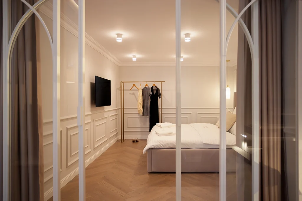 Serene bedroom framed by decorative arched glass partition, featuring crisp white linens, tailored paneling, and curated wardrobe accents in a minimalist architectural setting.