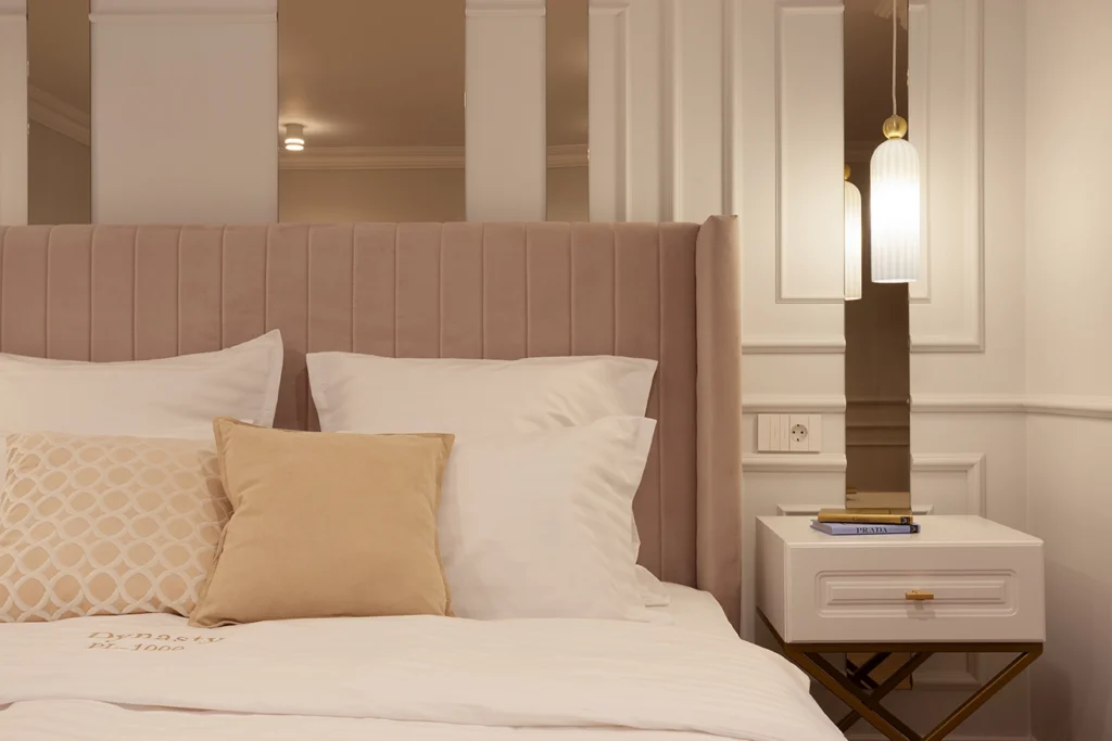 Modern bedroom with upholstered headboard, white linens, gold-accented nightstand, ribbed glass pendant lamp, and paneled wall detailing.