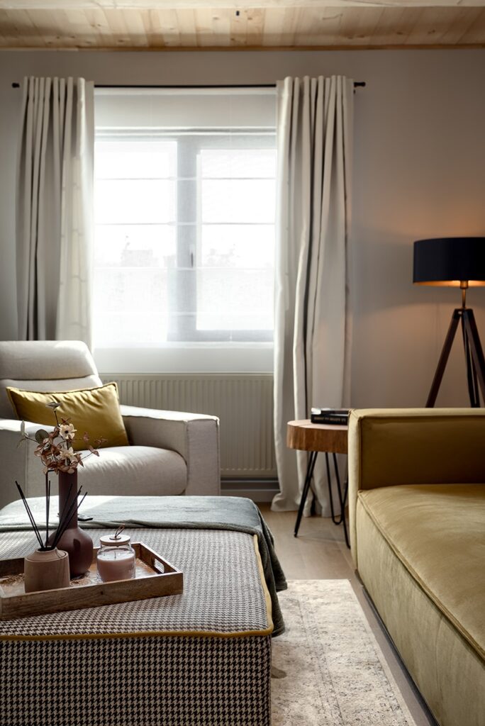 Cozy living room with olive green sofa, white armchair, woven ottoman coffee table, wood-paneled ceiling, and natural light from large window.