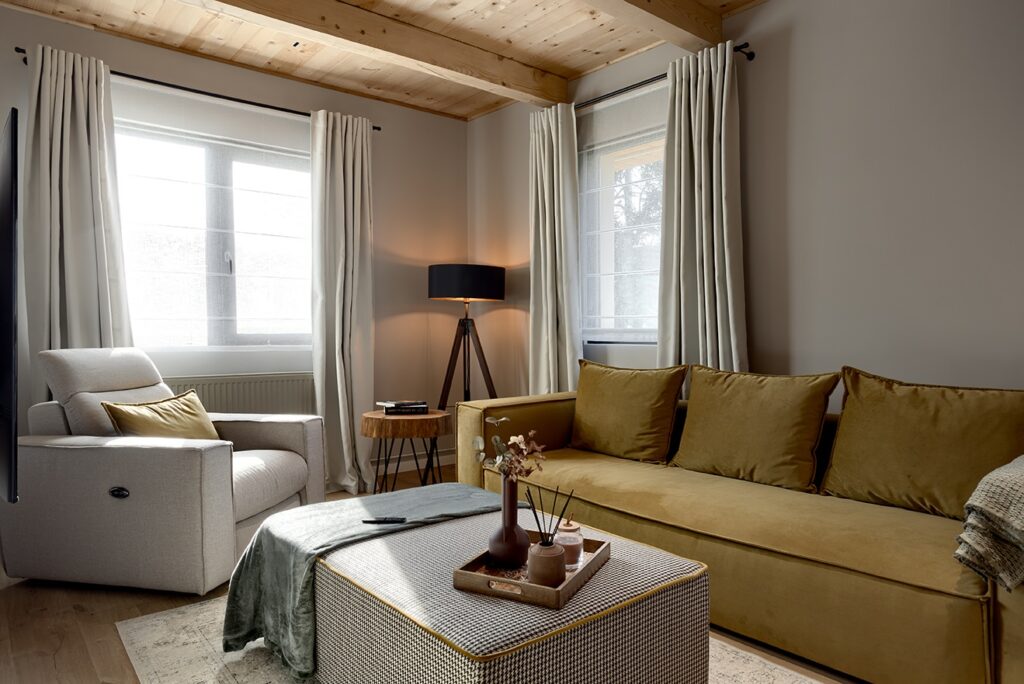 Cozy nordic living room with mustard sofa, gray recliner, stup side coffee table, checkered ottoman, exposed wood beam ceiling, and natural light from large windows.