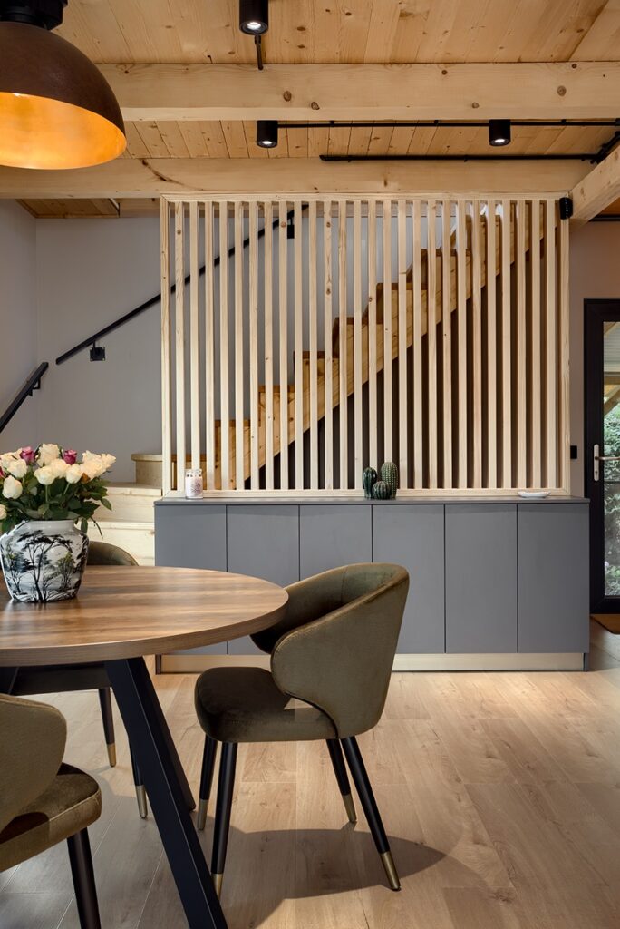 Modern dining area with round wooden table, velvet chairs, floral vase, and a staircase separated by vertical wooden slats and grey cabinetry.