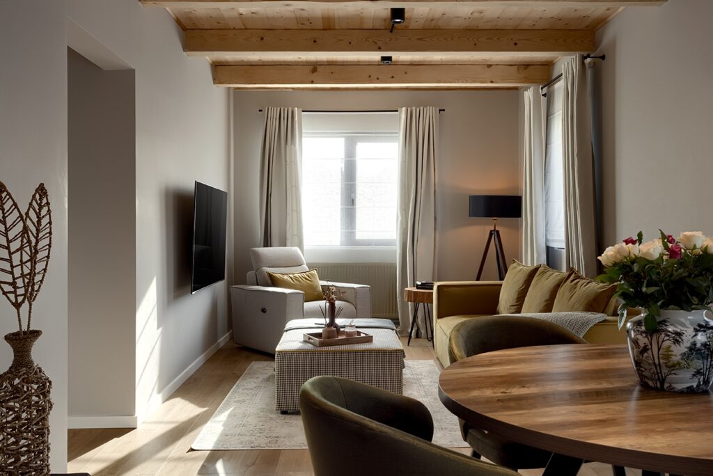 Modern cottage rustic living space with exposed wood beams, light walls, natural textures, and cozy furniture arranged around a warm, sunlit interior. Green velvet sofa, white armchair.