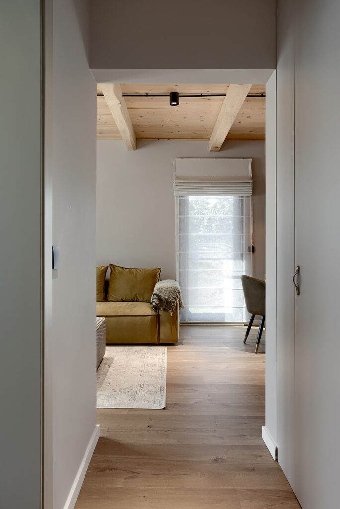 View from hallway into a warm modern living space with mustard yellow sofa, exposed wood beam ceiling, green accent chair, light area rug, and natural light through sheer curtains.