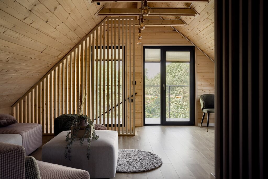Modern attic interior with wood-paneled walls and ceiling, exposed beams, hanging lights, glass doors to balcony, and cozy seating with natural textures