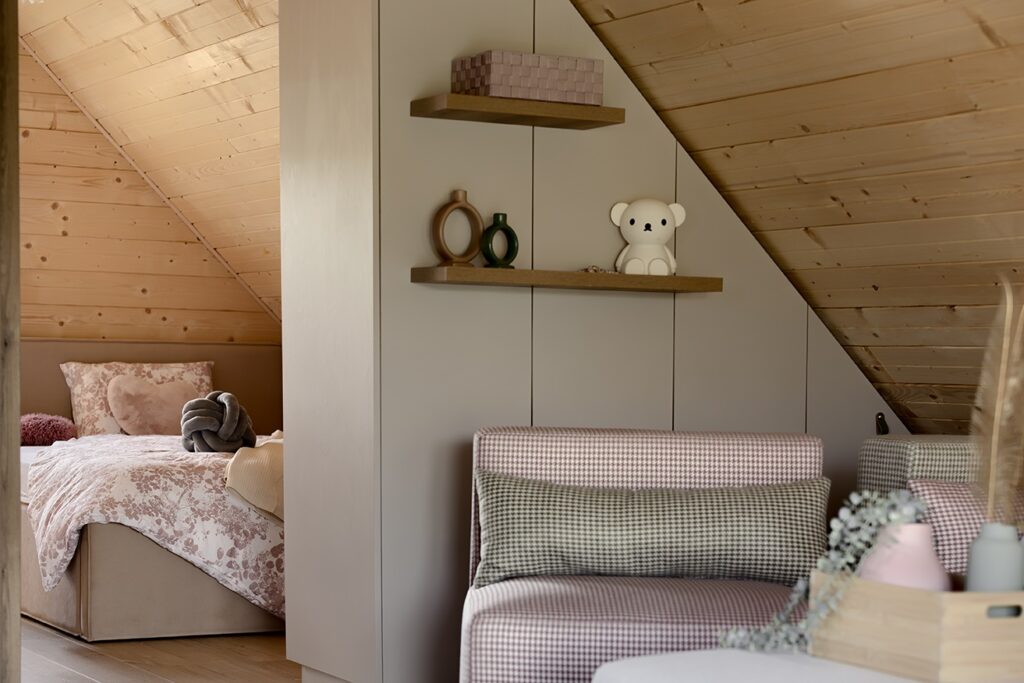 cozy bedroom with slanted wood-paneled ceiling, floral bedding, pastel seating area, and decorative shelves with soft toys, bear decoration, and geometric accents.