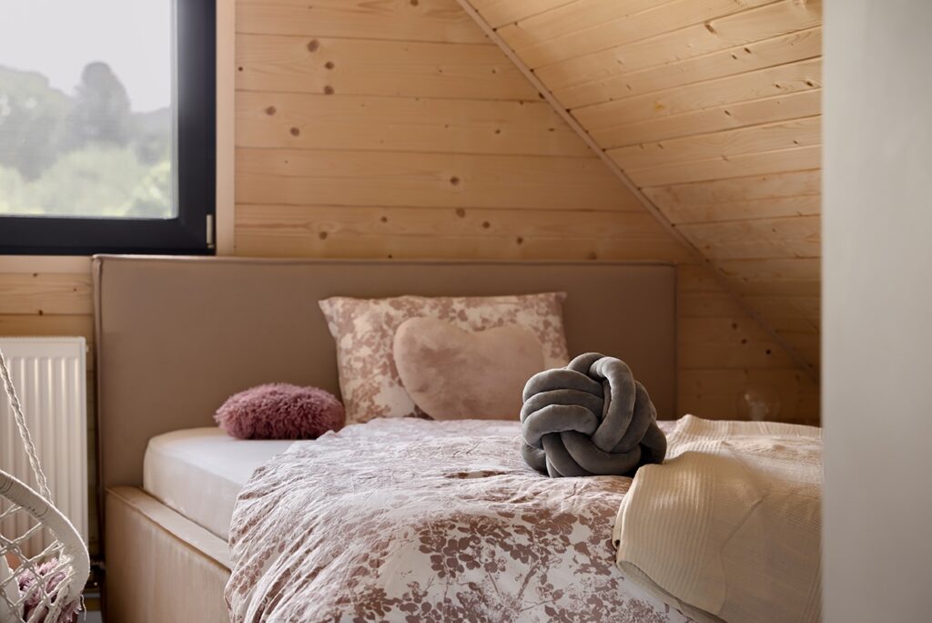 Charming kids’ attic bedroom with floral bedding neatly arranged on a low bed, featuring playful cushions including a gray knot pillow, heart-shaped accent, and fluffy round decor, all under a sloped wood-paneled ceiling with natural light from a black-framed window.