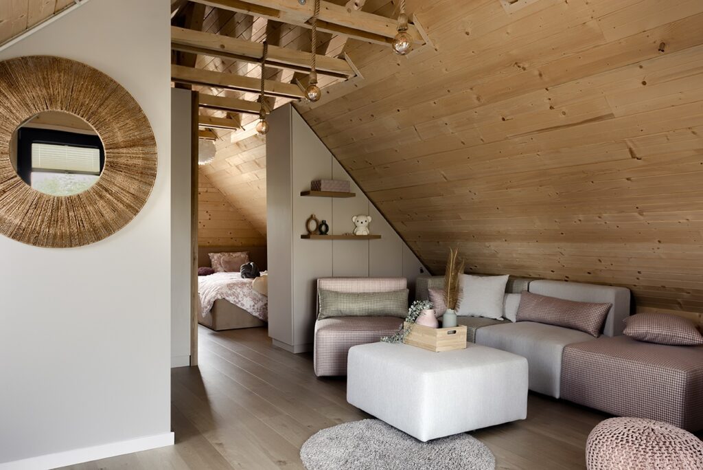 Warm attic living space with sloped wood ceiling, pink and gray sectional sofa, round woven-frame mirror, and cozy bedroom nook with patterned bedding.