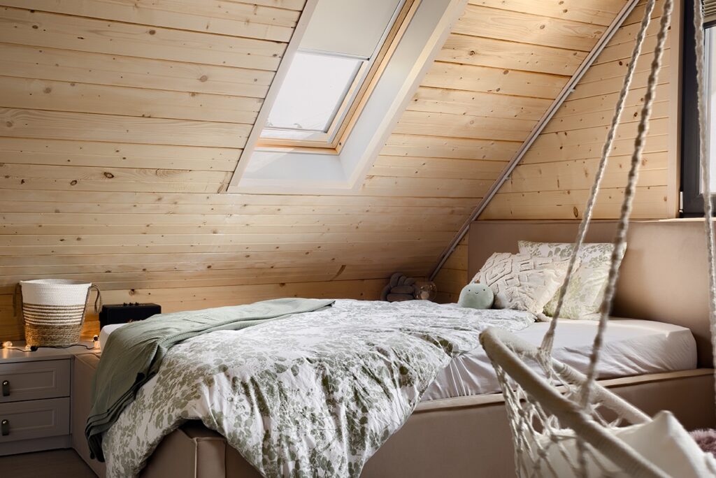 Rustic attic bedroom with sloped wood ceiling, skylight window, white and green bedding, knot pillow, and cozy nightstand with basket, Marshall speaker and string lights.