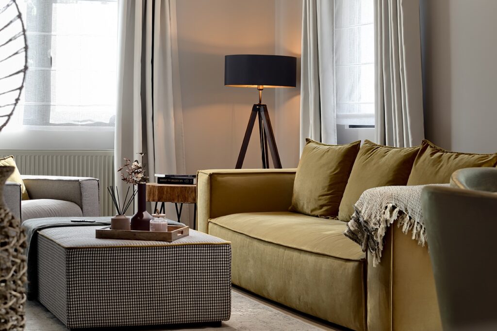 Contemporary living room with mustard yellow velvet sofa, houndstooth ottoman coffee table, gray armchair, and natural light from sheer-curtained windows.