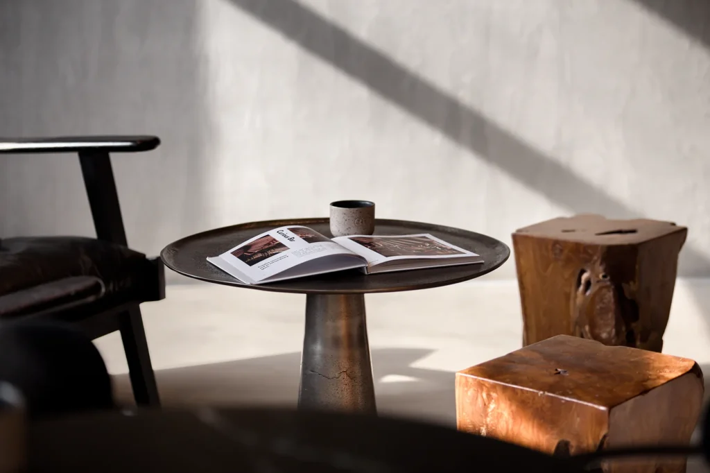 Minimalist lounge scene with round metal coffee table, open magazine, and ceramic cup, framed by sculptural wood stools and a leather-cushioned armchair. Diagonal natural light casts soft shadows across pale walls, emphasizing material contrast and spatial calm. Ideal for editorial storytelling in residential or hospitality design photography