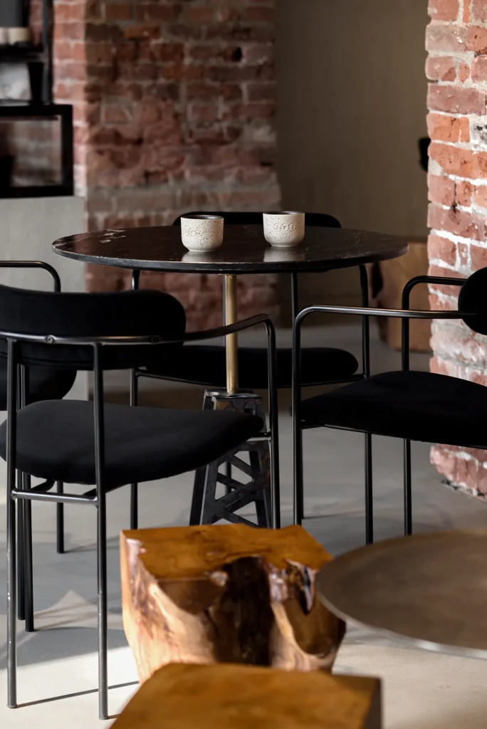 Cozy lounge vignette with black cushioned chairs around a round table, set against exposed brick walls and accented by a sculptural wood stool. Ceramic cups and soft textures evoke a tactile, intimate atmosphere. The interplay of raw materials and curated furniture highlights spatial warmth and editorial potential in hospitality design