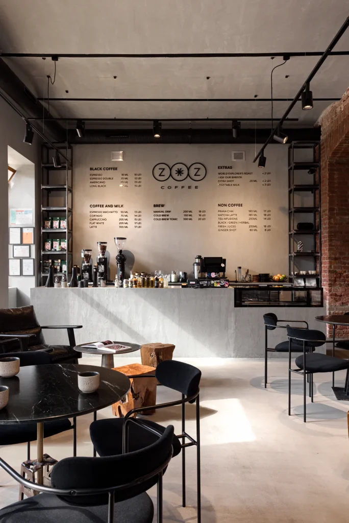 Raw-contemporary coffee shop interior with concrete surfaces, exposed brick, and matte black metal accents. The layout features intimate table clusters, visible espresso gear, and a branded menu wall that anchors the barista zone. The interplay of industrial restraint and curated hospitality branding makes this space ideal for commercial design storytelling and architectural photography