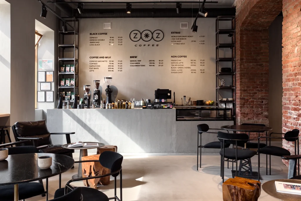Contemporary coffee shop interior with exposed brick, industrial lighting, and minimalist black furniture. The barista counter anchors the space with visible grinders, espresso machines, and a wall-mounted menu showcasing specialty brews and milk alternatives. The spatial clarity and material contrast reflect a brand-conscious hospitality design—ideal for editorial documentation and commercial portfolio work