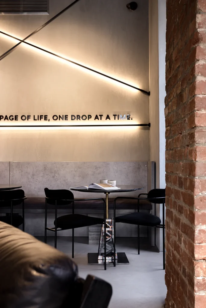 Minimalist café lounge with illuminated quote wall, sculptural lighting, and curated seating vignette featuring black chairs, round table, and open book with coffee cup. Brick textures and leather accents frame the scene, blending tactile warmth with editorial clarity. Captures brand storytelling and spatial intimacy in hospitality design shot by Ilisie Sergiu Adrian interior photographer