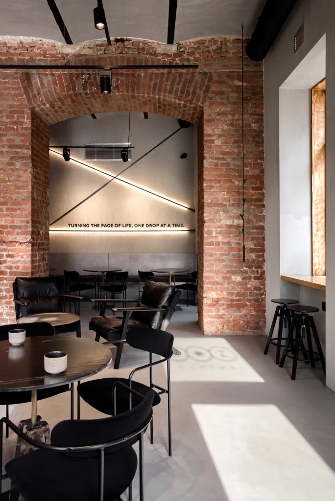Stylized café interior blending raw materials and refined geometry—arched brickwork, polished concrete floors, and sculptural black furniture. The space features layered seating zones, a bar-height counter near natural light, and diagonal wall lighting that frames a poetic quote. Captures spatial rhythm and brand storytelling in hospitality design