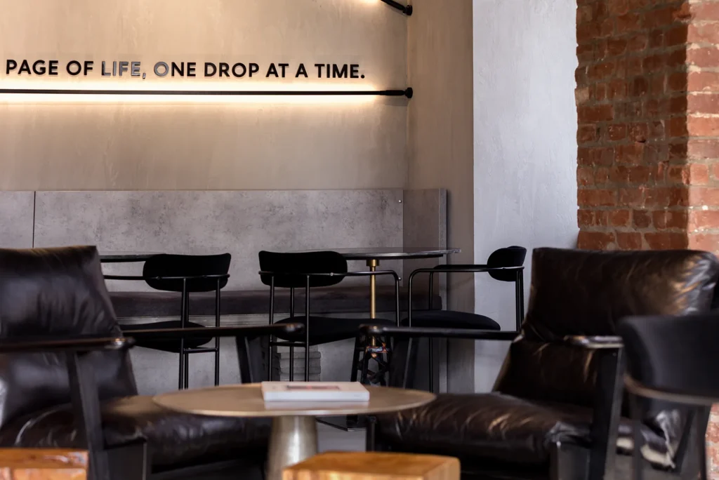 Modern café interior with exposed brick walls, concrete textures, and warm ambient lighting. Features include black leather armchairs, minimalist black chairs, and a stone table with integrated wall lighting that highlights motivational typography. The industrial-contemporary blend and curated seating layout showcase a hospitality space ideal for editorial and commercial design photography