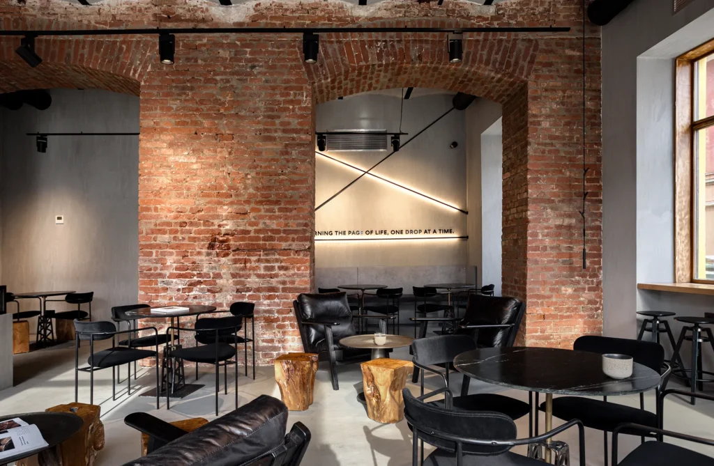 Industrial-style café interior with exposed brick arches, concrete walls, and minimalist black furniture. Features include round tables, leather armchairs, rustic wood stump stools, and diagonal wall lighting. The bold typography on the back wall and curated textures highlight a visually compelling hospitality space—ideal for editorial, branding, and commercial design portfolios