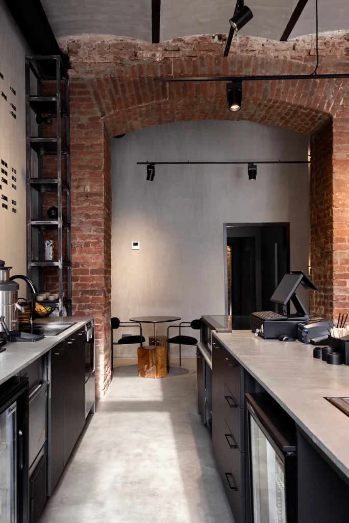 Industrial café interior with arched brick ceiling, concrete countertops, and matte black cabinetry. The narrow layout integrates barista equipment, open shelving, and a rear seating vignette with glass table, sculptural wood accent, and black chairs. Track lighting and material layering emphasize depth, rhythm, and operational flow—ideal for documenting spatial choreography in hospitality design