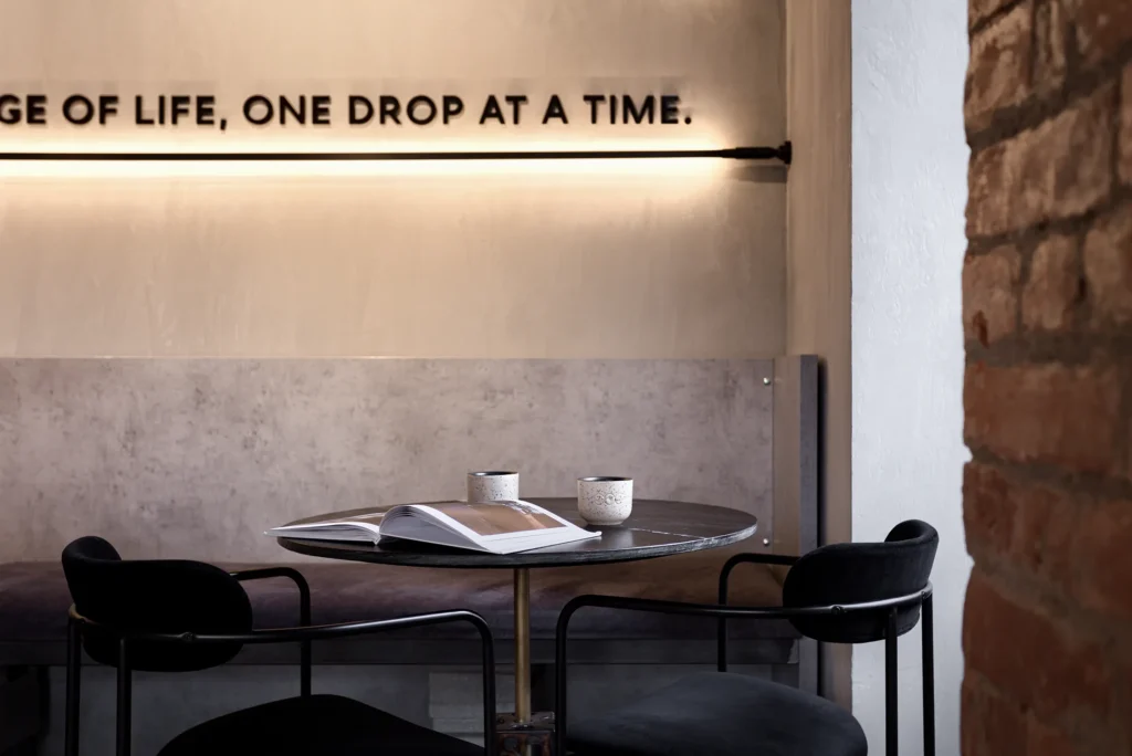 Modern café interior with round black table, brass pedestal base, and four upholstered chairs in matte black metal. The scene includes ceramic cups and an open magazine, framed by a concrete wall with integrated lighting and bold typography. Brick accents and layered textures support a brand-forward hospitality concept ideal for editorial, social media, and commercial design photography portfolios
