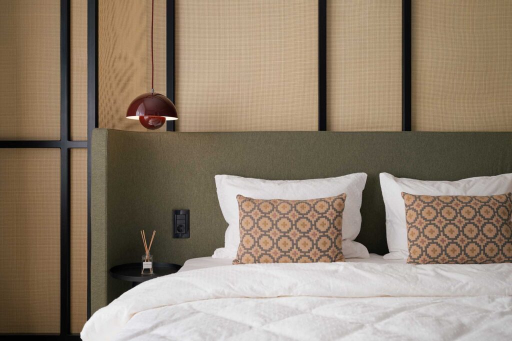 Close‑up interior magazine photo bedroom with green upholstered headboard, geometric wall panels in beige and black, and a brown dome pendant light, featuring a decorative pillow with woven geometric texture in beige and terracotta tones, white bedding, and soft beige throw.