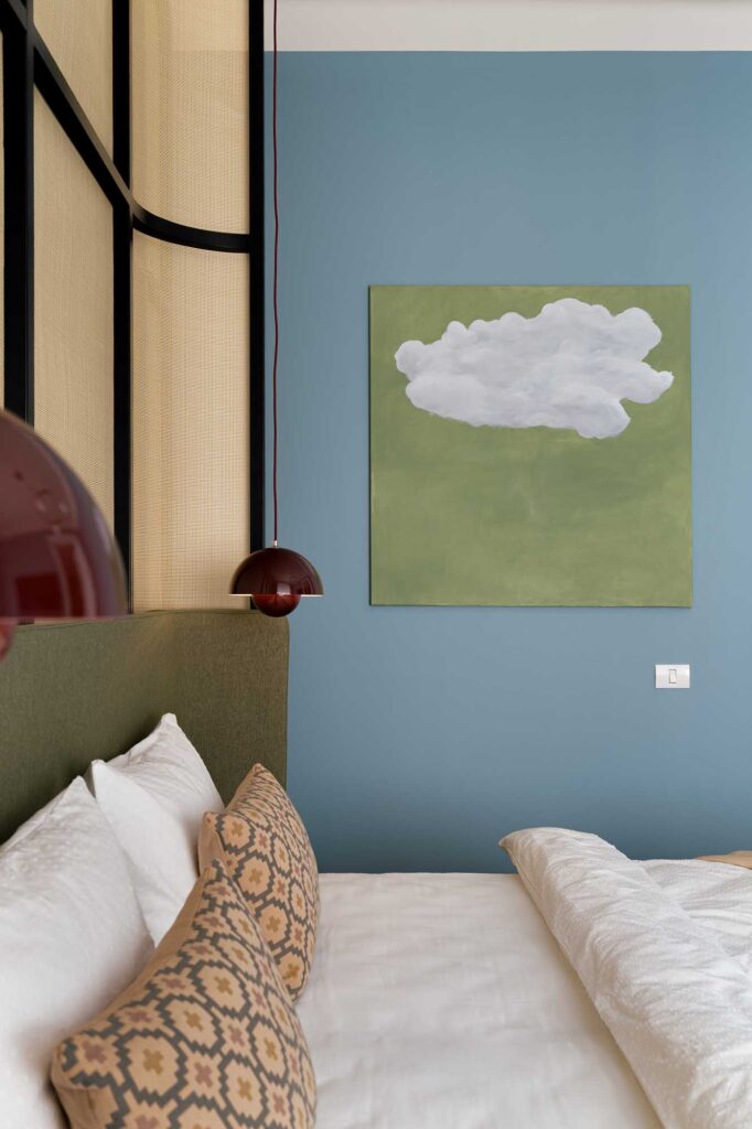 bedroom with green upholstered headboard, patterned beige and brown woven cushions, and minimalist wall art depicting a white cloud on green background painted by the designer Madalina Nini, complemented by blue accent wall, red dome pendant light, and soft white bedding.