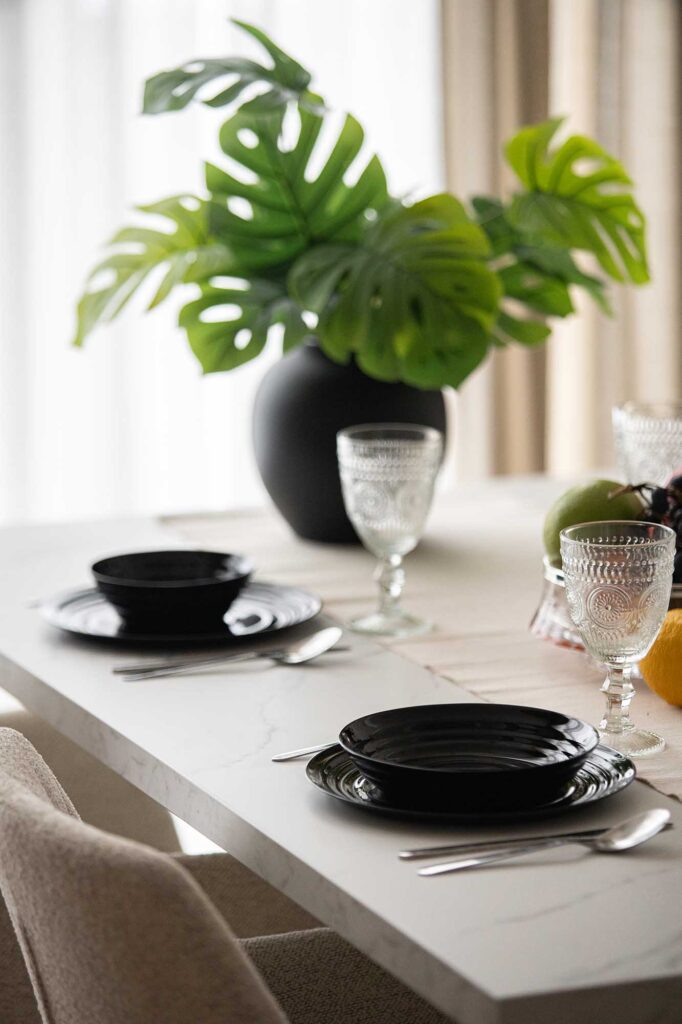 Elegant hospitality dining setup featuring a marble‑look table styled with black ceramic plates, crystal glasses, and a matte black vase holding lush monstera leaves — a refined composition that blends natural freshness with modern sophistication, ideal for boutique hotels and short rental apartments.