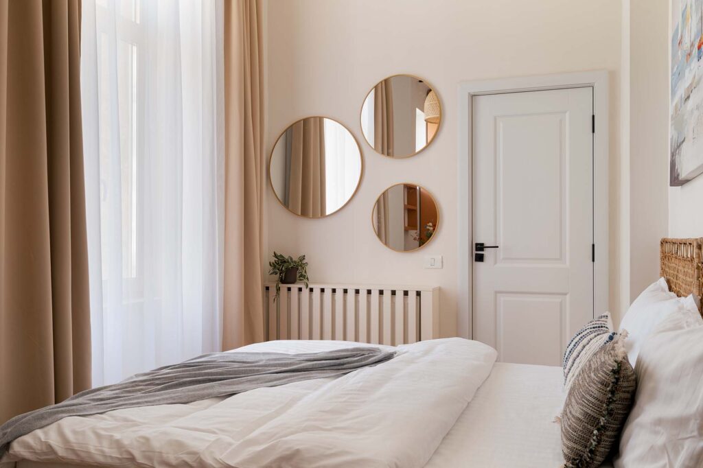 Bright modern, cozy and calm bedroom with white bedding, woven headboard, and round wall mirrors reflecting natural light, complemented by beige curtains, soft cream walls, and minimalist decor with a small potted plant on radiator for a serene atmosphere.