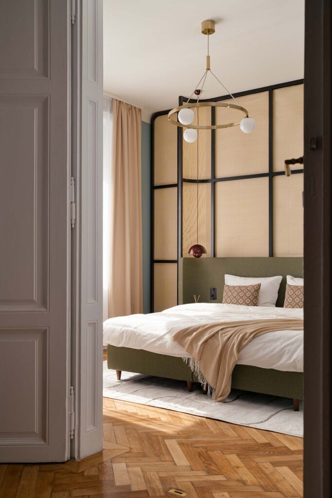 Contemporary bedroom with green upholstered bed, beige throw blanket, and geometric panel wall featuring woven textures and black framing, complemented by brass pendant light, herringbone parquet flooring, and soft natural light through beige curtains.