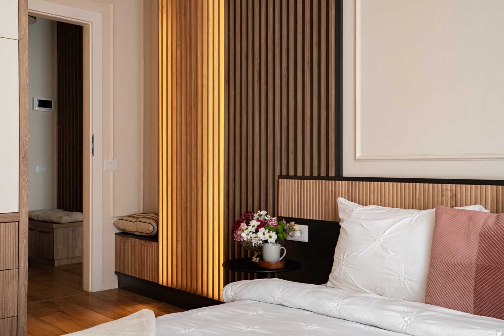 Editorial hospitality photograph showcasing a boutique bedroom with vertical wood slats illuminated by warm ambient light, crisp white bedding, blush accent pillow, and floral arrangement, capturing the essence of crafted comfort and refined atmosphere for modern hotels and guest experiences.