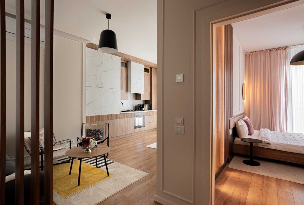 Hospitality apartment interior featuring open‑plan living area with light wood flooring, marble kitchen cabinetry, and cozy bedroom framed by soft pink curtains — captured in natural light to highlight warm wood textures, refined materials, and seamless spatial flow for boutique accommodations and serviced apartments. Photographed by Sergiu Ilisie
