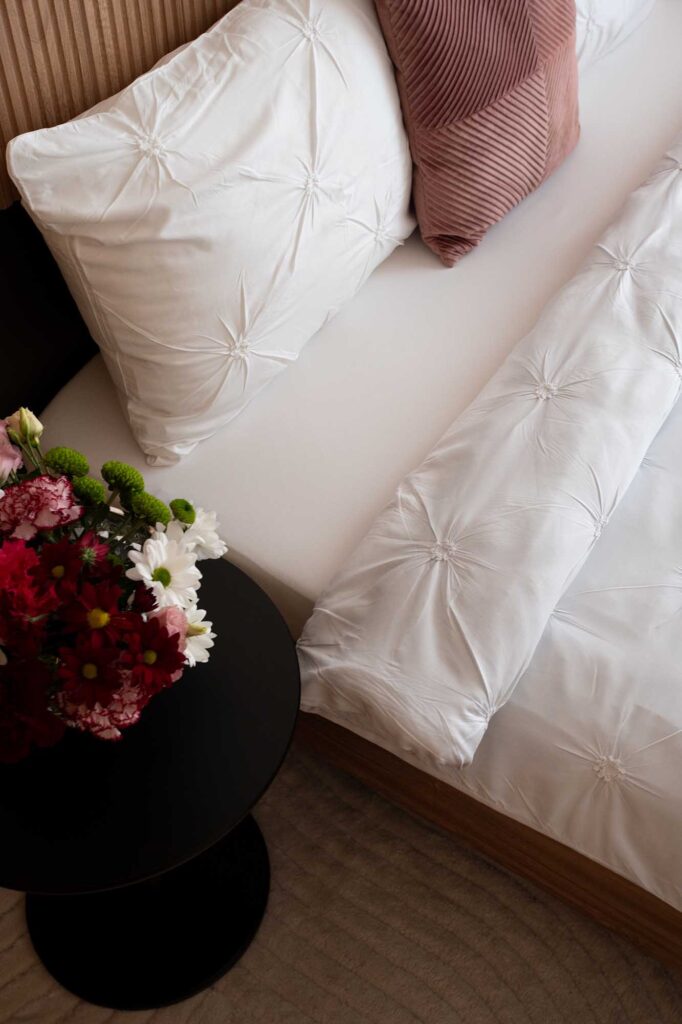 Hospitality‑style bedroom detail featuring textured white bedding, ribbed rose pillow, and a round black bedside table with a vibrant floral arrangement, creating a warm and inviting atmosphere ideal for boutique hotels and modern guest accommodations