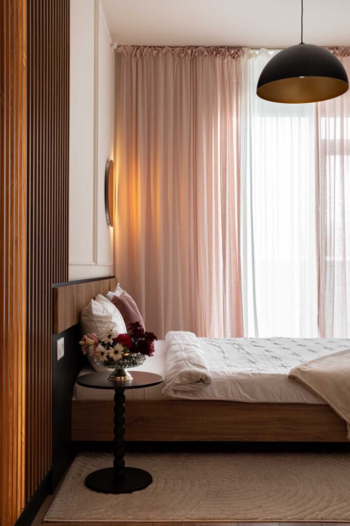 Hospitality‑style bedroom interior featuring soft pink curtains, warm ambient lighting, and a modern wooden bed with textured bedding and floral arrangement, captured in natural light to highlight comfort and elegance for boutique hotels and guest accommodations