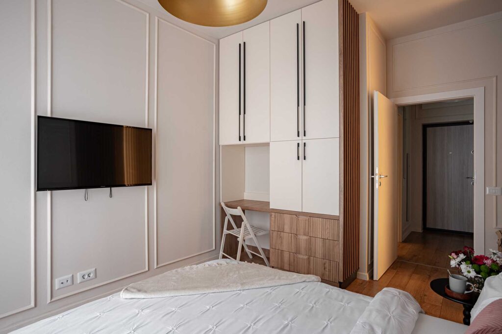 Hospitality interior photograph of a modern bedroom featuring white quilted bedding, built‑in wardrobe with black handles, and compact workspace with folding chair, captured in soft daylight to highlight functional elegance and refined design for boutique hotels and serviced apartments