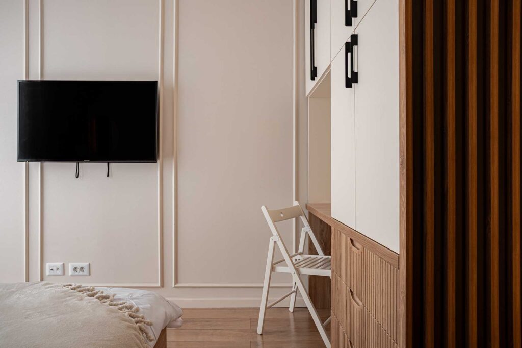 Hospitality‑style bedroom interior featuring minimalist design with wall‑mounted TV, white folding chair, and built‑in wooden cabinetry with black handles, captured in soft natural light to highlight clean lines and modern comfort for boutique hotels and guest accommodations