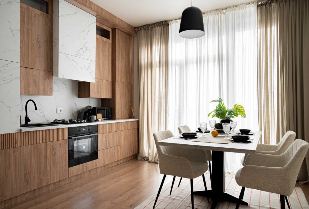 Hospitality kitchen and dining area featuring natural wood cabinetry with marble accents, a white dining table set with black tableware, and soft beige curtains filtering daylight — a refined composition that blends warmth, texture, and modern elegance for boutique hotels and short-rental apartments