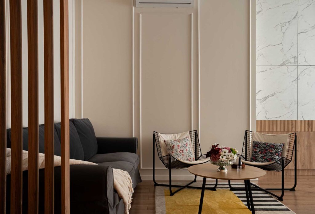 Hospitality living area featuring dark gray sofa, floral lounge chairs, and round wooden coffee table with fresh flowers, complemented by marble MDF cabinetry and warm wood accents, captured in natural light to highlight contemporary design and inviting atmosphere for boutique hotels and serviced apartments