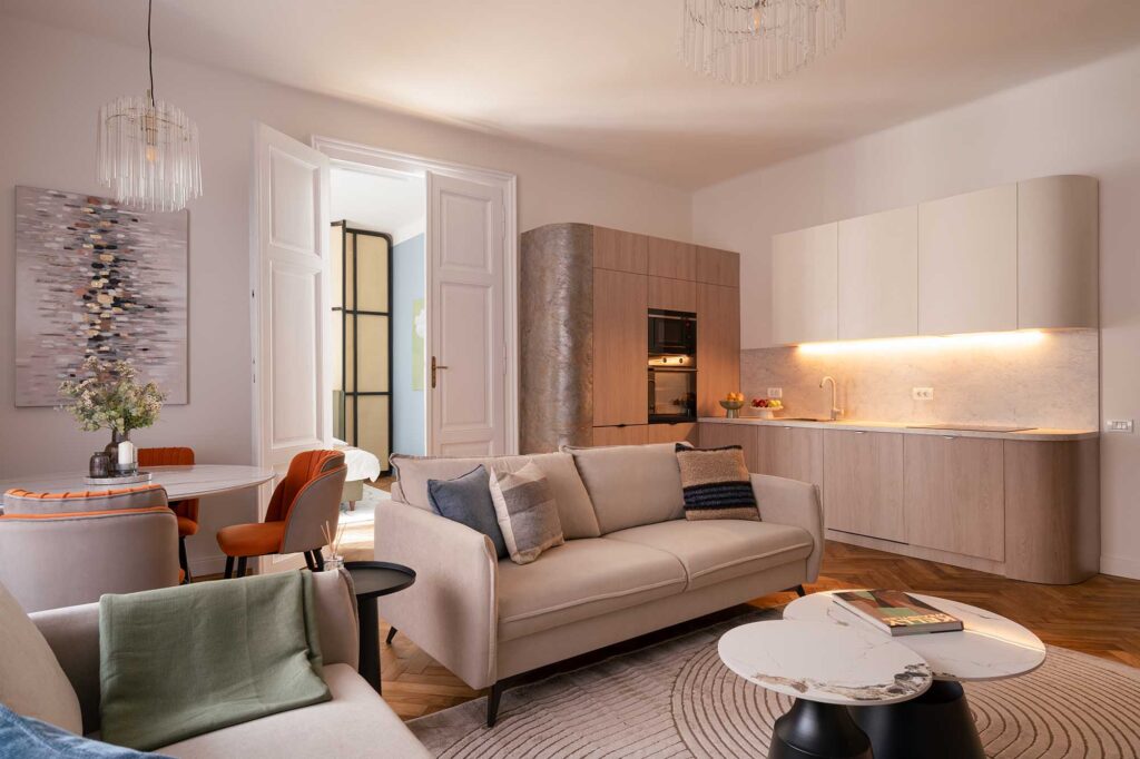 Stylish open‑space living space with beige sofas, marble coffee tables, and integrated kitchen featuring light wood cabinetry, stone backsplash, and warm under‑cabinet lighting, complemented by orange dining chairs and abstract wall art.