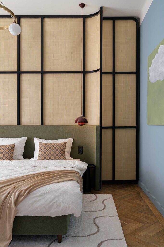 Editorial photography captured by ILI HAUS Ilisie Sergiu with a contemporary bedroom with green upholstered bed, beige throw blanket, and geometric wall panels featuring woven textures and black framing, complemented by brass pendant light, herringbone parquet flooring, and minimalist artwork on blue wall.
