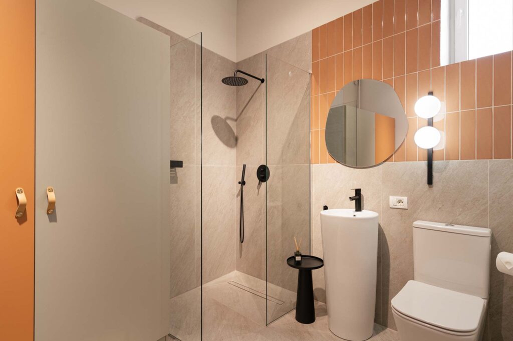 Contemporary bathroom with terracotta vertical tiles, light stone walls, and glass shower enclosure featuring matte black fixtures, round mirror, sculptural white pedestal sink, and modern wall light, complemented by warm orange cabinetry and minimalist design.
