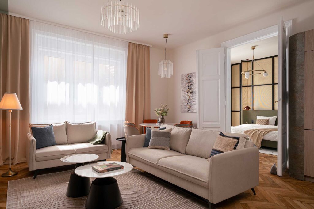 Architectural Photography of an elegant modern apartment interior with beige sofas, marble coffee tables, designer chandeliers, and open view into bedroom with geometric wall panels and brass pendant light.