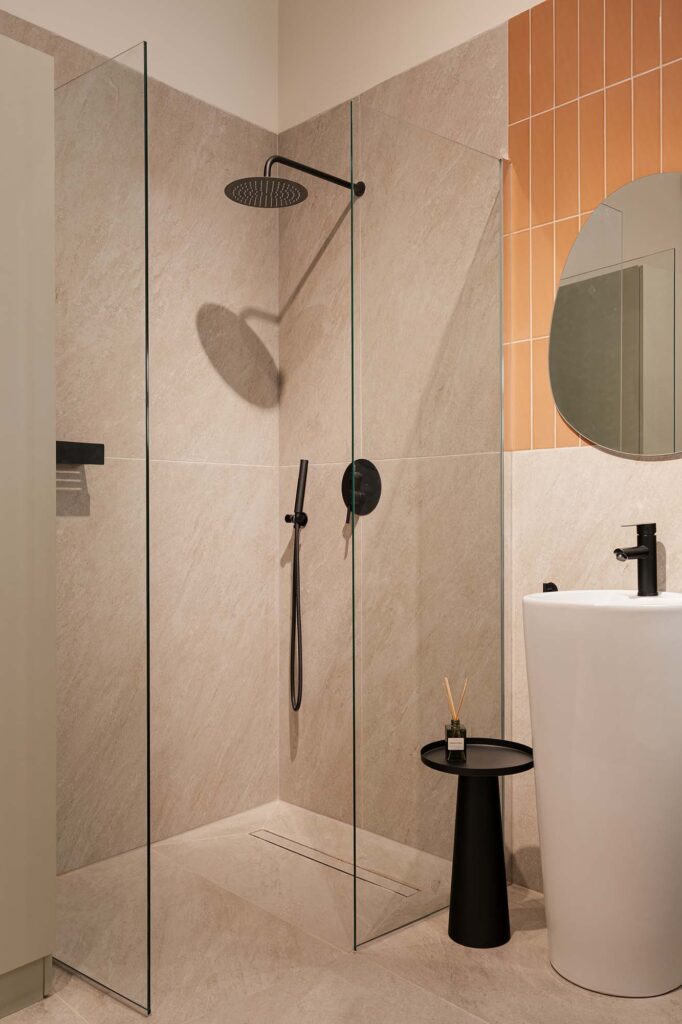 Minimalist bathroom with terracotta vertical tiles and beige stone walls, featuring a glass shower enclosure with matte black rain shower and handheld fixtures, white pedestal sink with black faucet, and small black side table with reed diffuser for a warm, contemporary aesthetic.