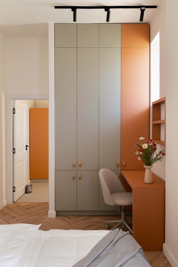 Modern bedroom corner featuring tall built‑in wardrobe in muted gray and burnt orange, matching desk with beige upholstered chair, and decorative vase with colorful flowers, complemented by herringbone wood flooring and soft natural light.